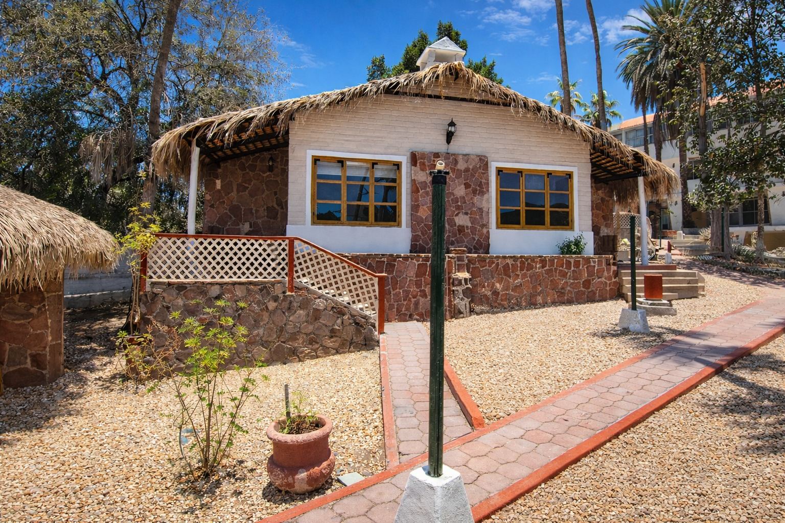 Cabaña con techo de palma, muros de piedra y ventanas de madera, rodeada de palmeras