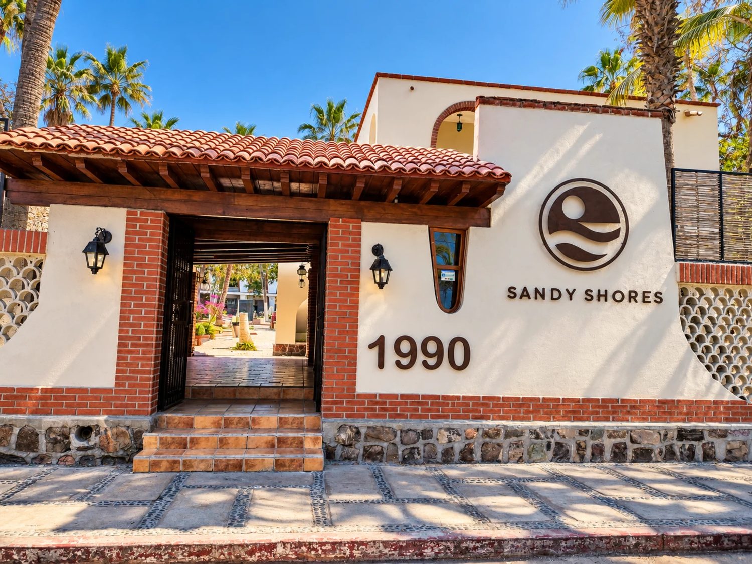Entrada de Sandy Shores: portón de herrería bajo un techo de teja, con el logo y el número 1990 en el muro