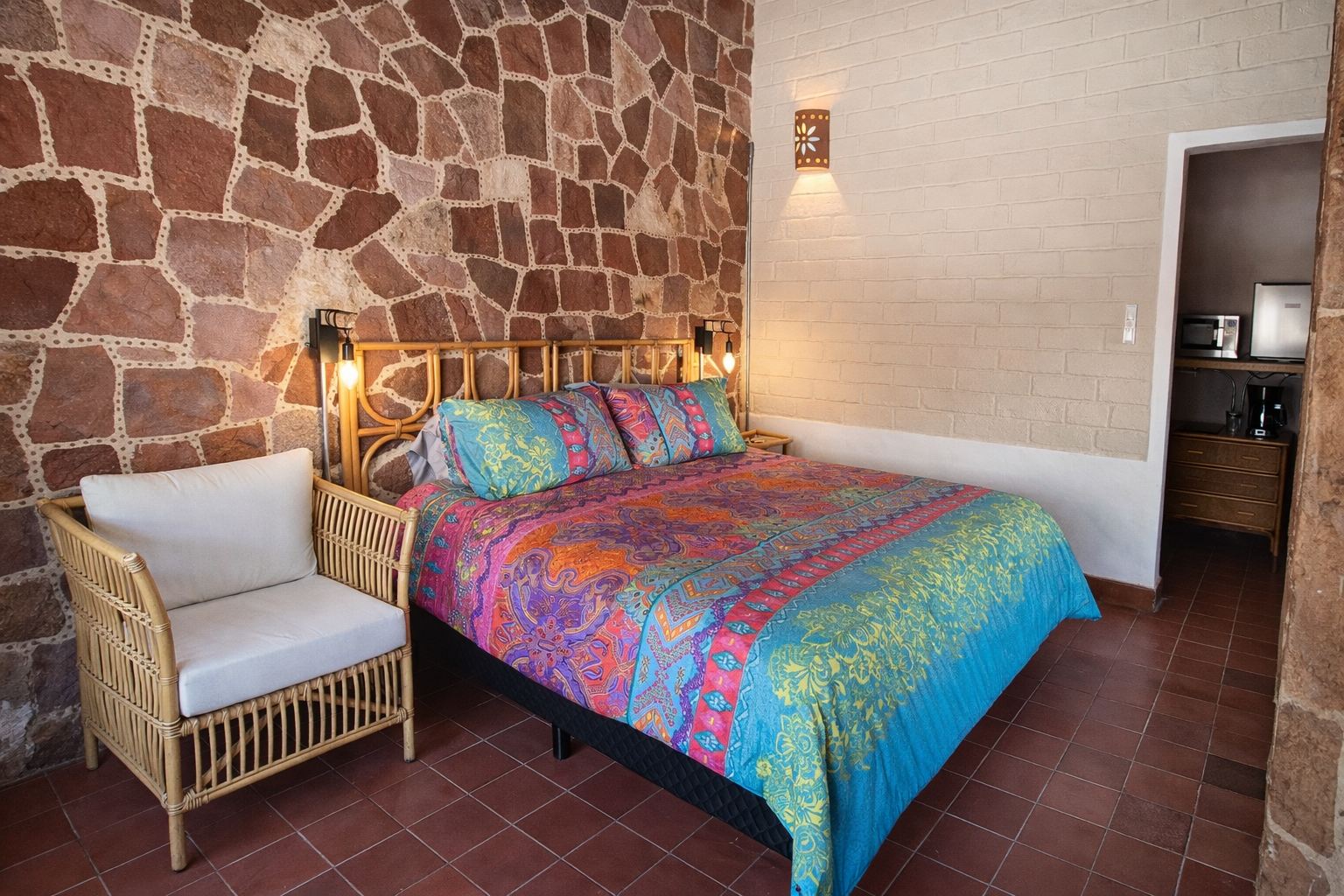 Cama King con cabecera de ratán frente a un muro de piedra, con sillón blanco y lámparas colgantes